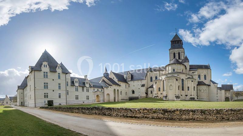 Vue panoramique d'une abbaye médiévale en pierre claire, entourée de pelouses et d'un chemin de terre sous un ciel…