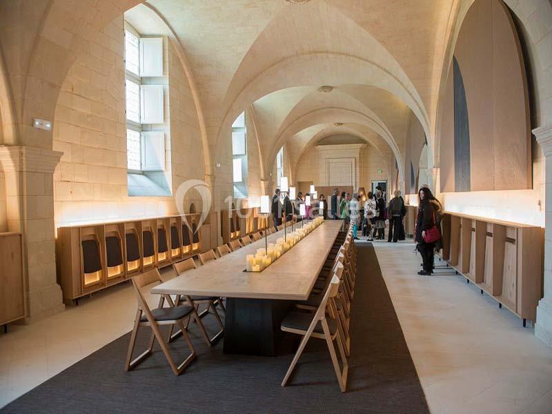 Salle voûtée avec une longue table en bois entourée de chaises, éclairée par des fenêtres et des luminaires discrets.