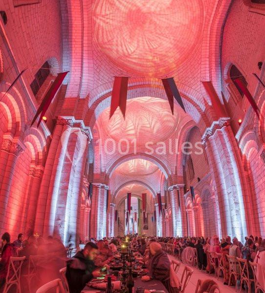 Salle voûtée illuminée de rouge avec de longues tables dressées pour un banquet, entourées de convives.