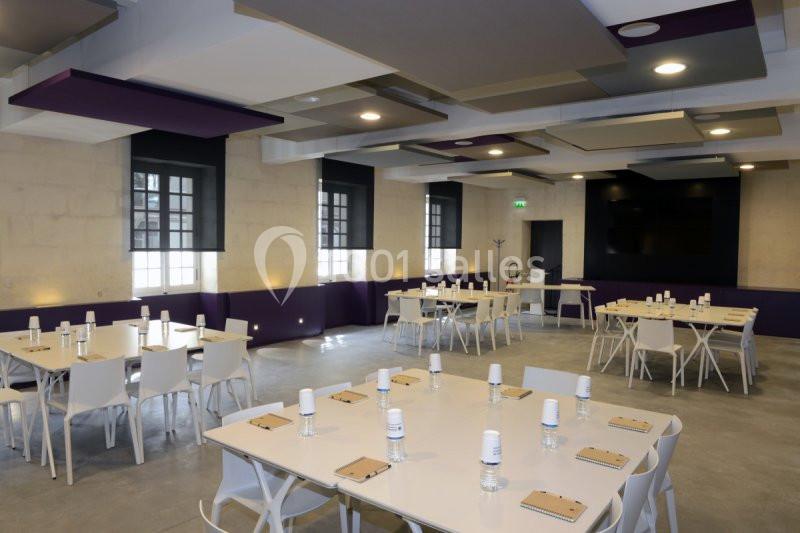 Salle lumineuse aménagée avec des tables blanches, chaises modernes et un plafond avec panneaux acoustiques colorés.