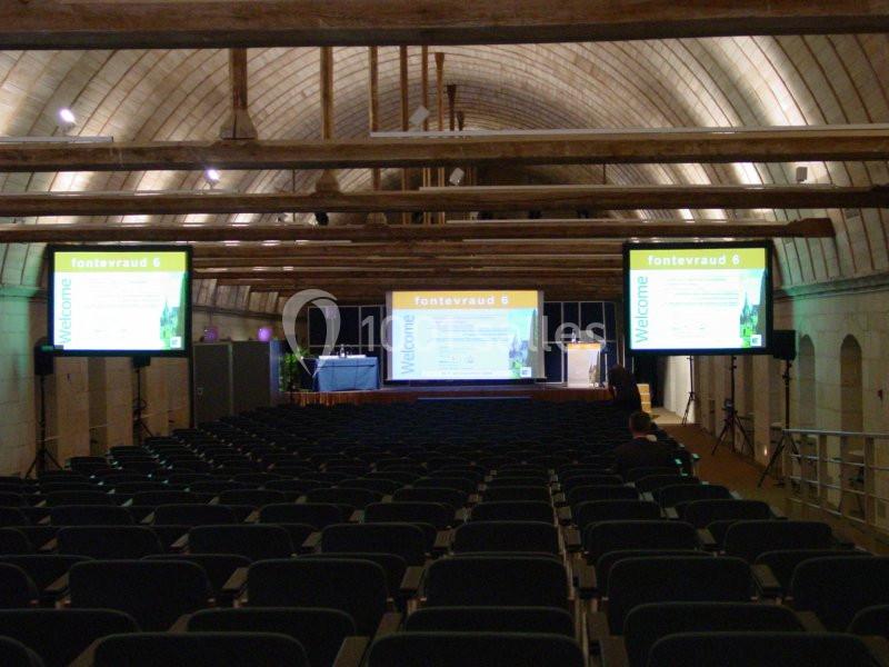 Salle de conférence voûtée avec rangées de sièges vides, écrans de présentation et estrade au fond.