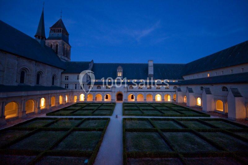 Vue nocturne d'un cloître avec jardins géométriques, éclairages intérieurs et une église en arrière-plan.