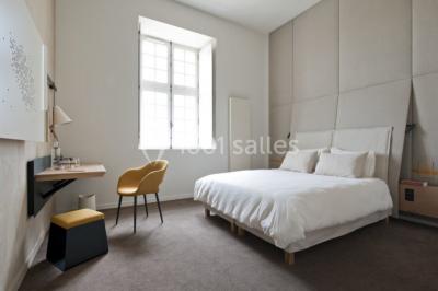 Chambre lumineuse avec lit double, mur beige capitonné, bureau minimaliste, chaise jaune et fenêtre à croisillons.