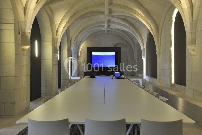 Salle de réunion avec une longue table blanche, des chaises, des voûtes en pierre et un écran allumé au fond.
