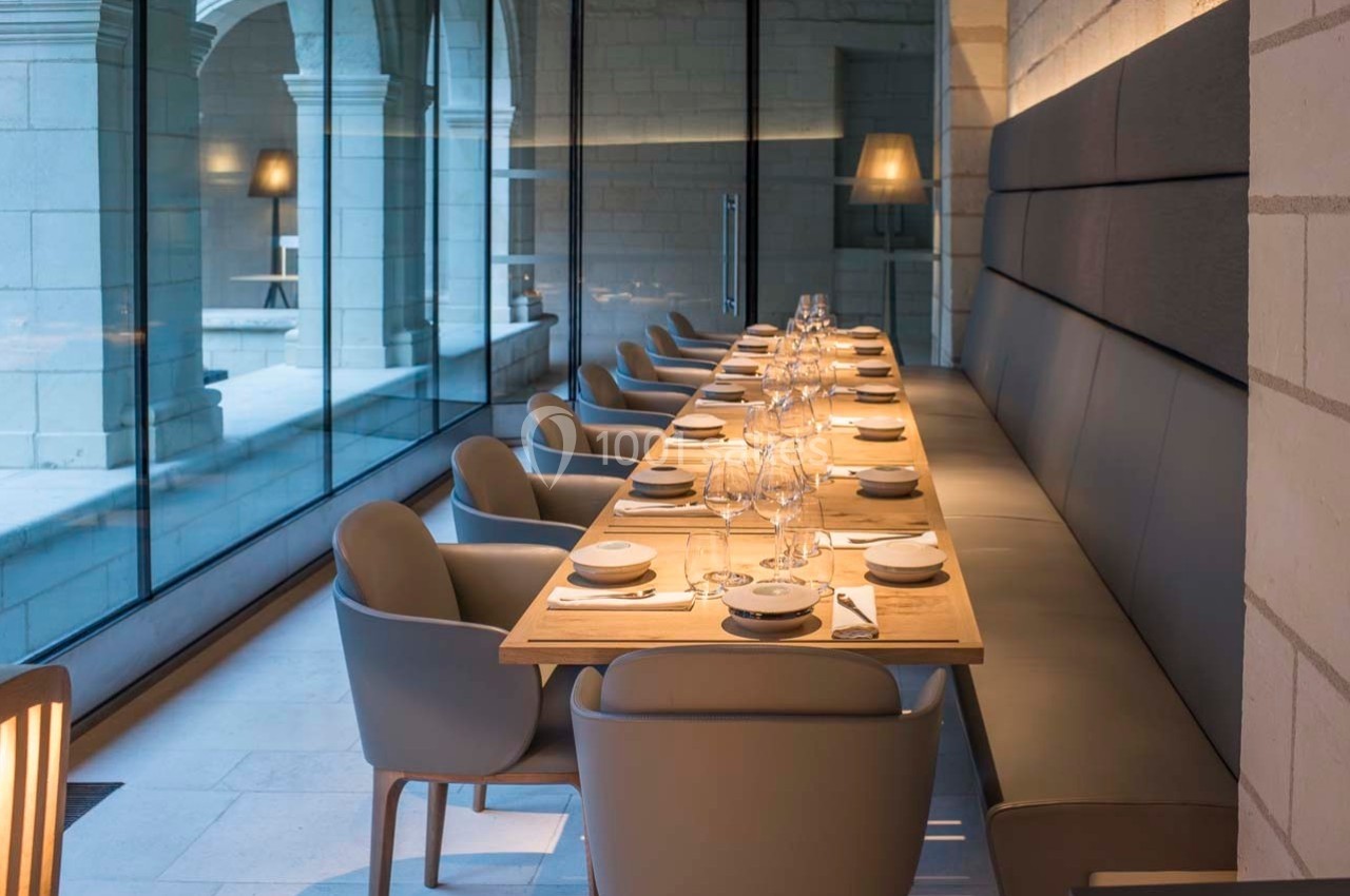 Salle de restaurant moderne avec une longue table dressée, chaises grises et grandes baies vitrées donnant sur une cour.