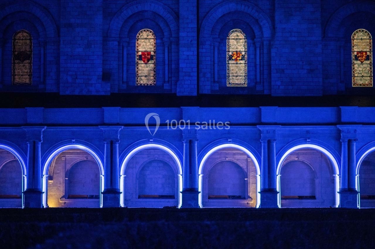 Façade d'un bâtiment en pierre avec des arches illuminées en bleu et des vitraux colorés visibles en hauteur.