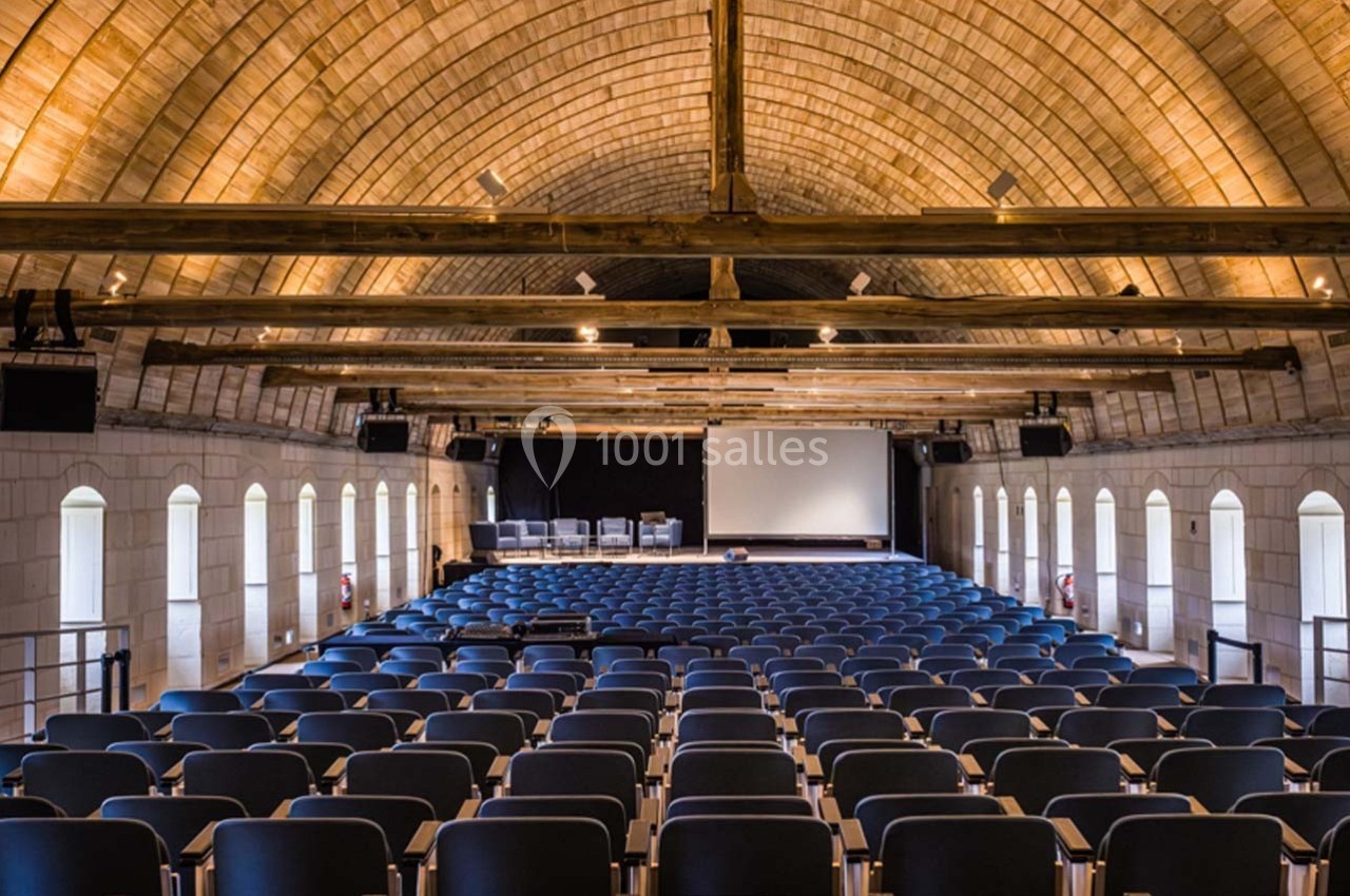 Salle de conférence avec rangées de sièges vides, écran de projection et plafond voûté en bois.