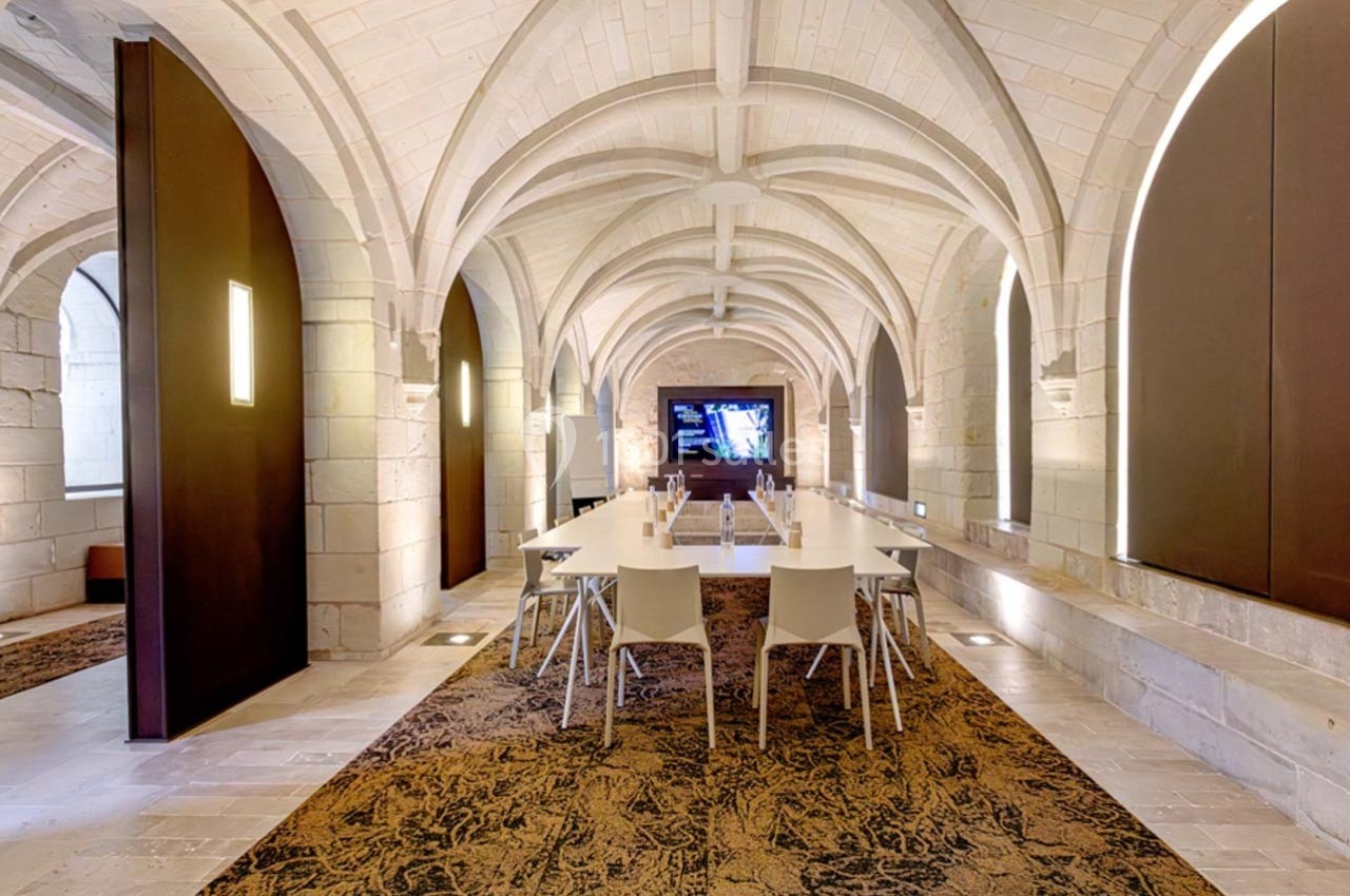 Salle voûtée en pierre avec une grande table blanche, des chaises modernes et un écran mural allumé.