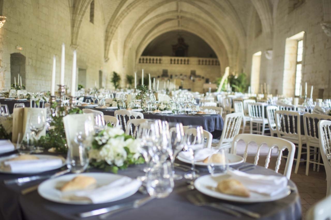 Salle voûtée décorée pour un événement, avec tables dressées, nappes grises, chandeliers et compositions florales.