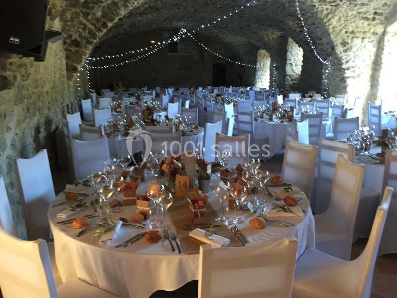 Salle voûtée en pierre aménagée pour un banquet, avec tables rondes dressées et chaises blanches.