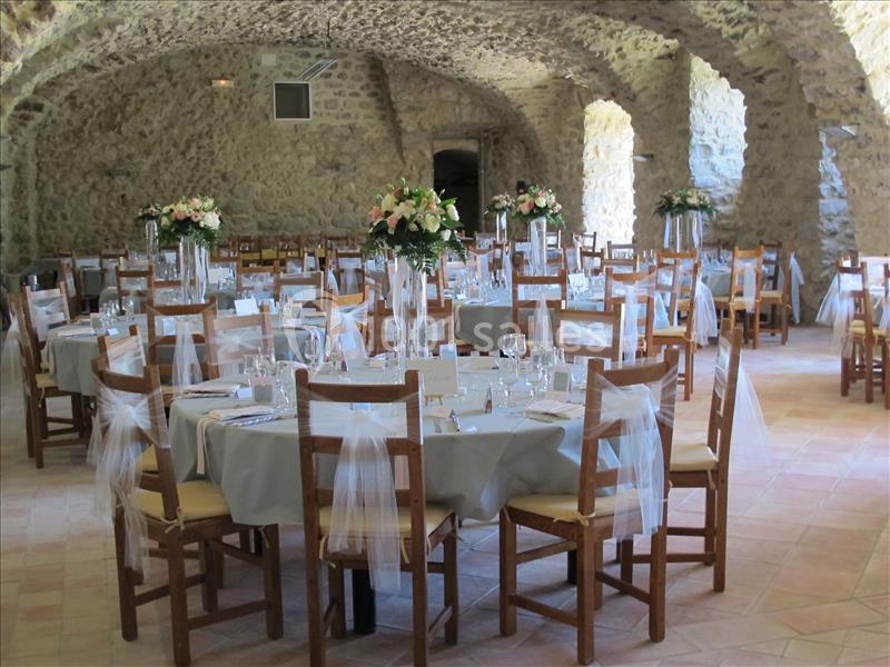 Salle voûtée en pierre aménagée pour un événement, avec tables rondes décorées de nappes, fleurs et chaises ornées de rubans.