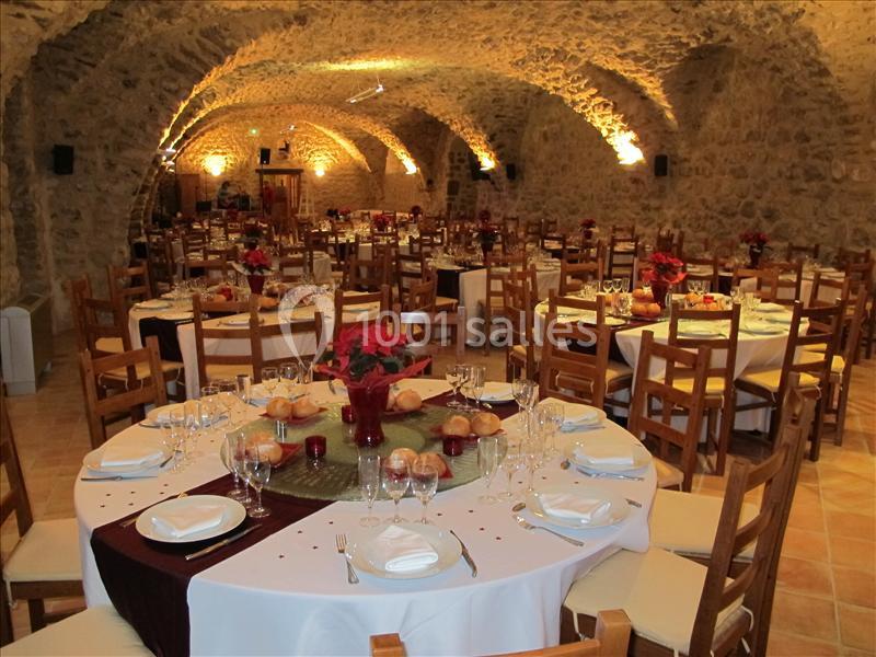 Salle voûtée en pierre aménagée pour un repas, avec tables rondes dressées, nappes blanches et décorations rouges.