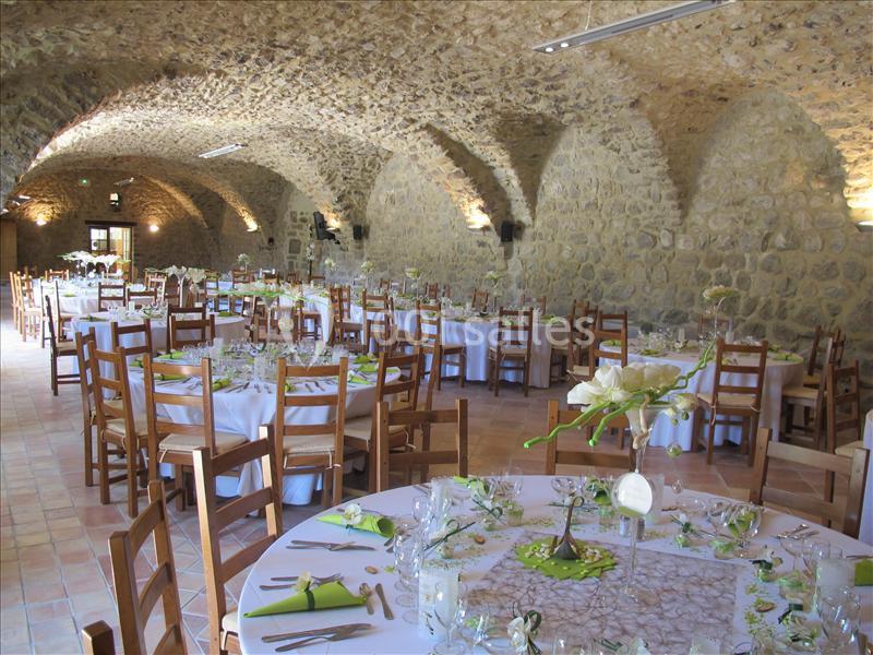 Salle voûtée en pierre aménagée pour un événement, avec des tables rondes décorées et des chaises en bois.