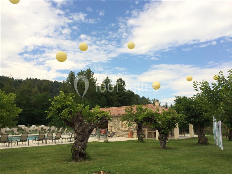 Maison en pierre avec toit en tuiles, entourée d'arbres, pelouse et piscine, sous un ciel décoré de ballons jaunes.