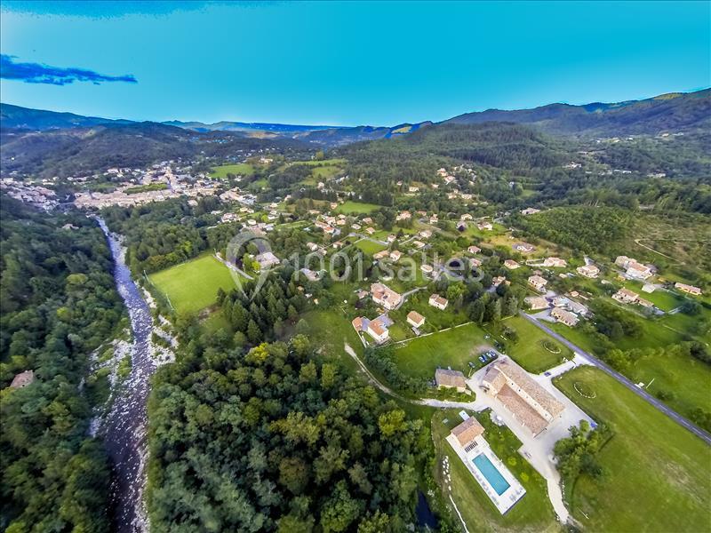 Vue aérienne d'un village entouré de collines verdoyantes, avec une rivière et des maisons dispersées.