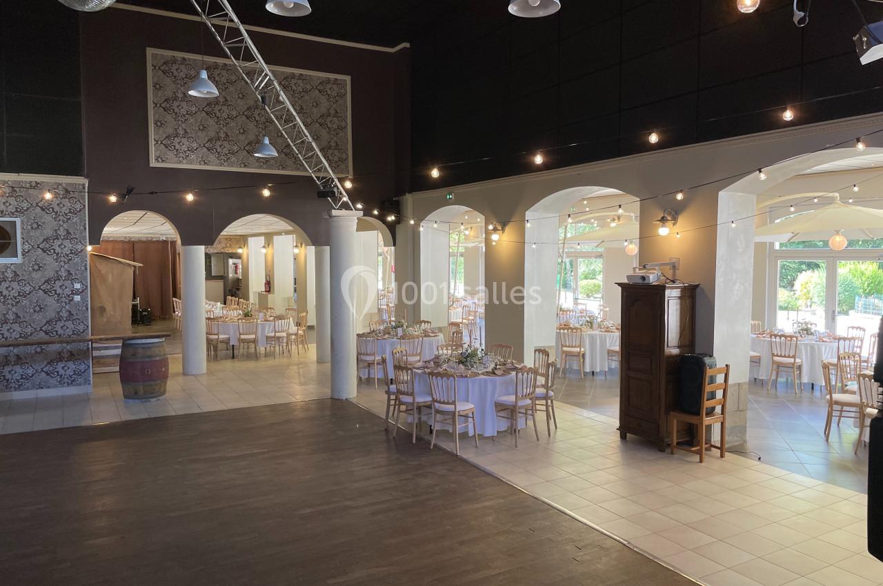 Salle de réception lumineuse avec tables dressées, chaises en bois et décorations élégantes sous des arches blanches.