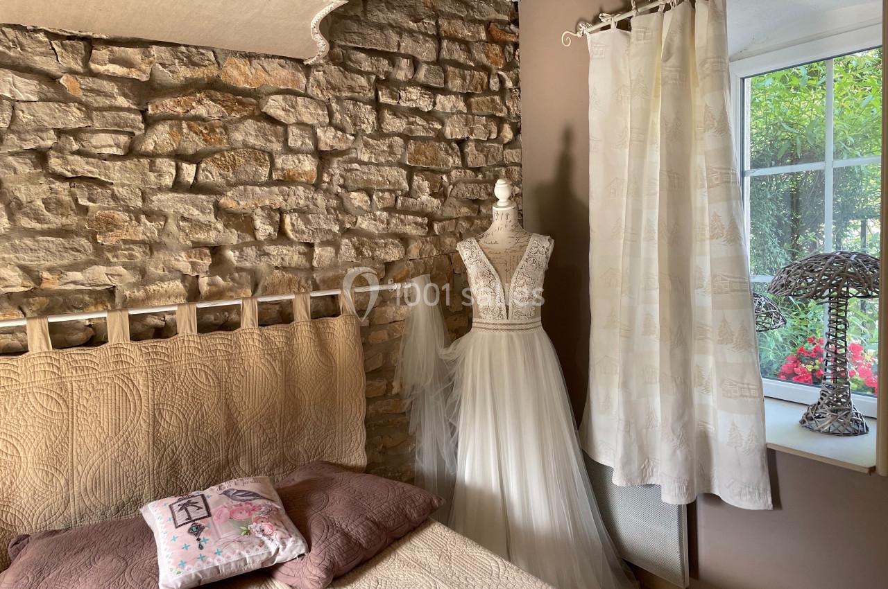 Mannequin avec robe de mariée en dentelle et tulle dans une chambre rustique avec mur en pierre et fenêtre donnant sur un…