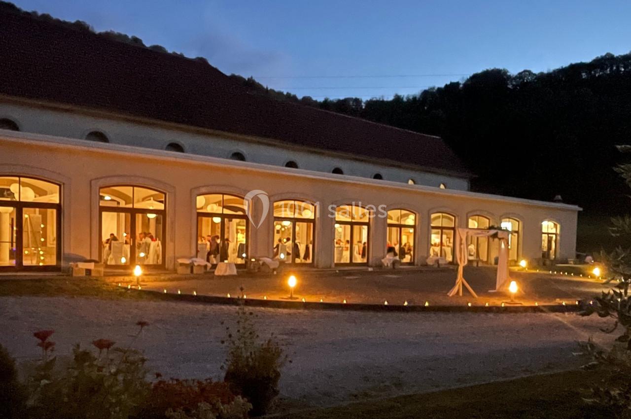 Salle de réception éclairée en soirée, avec de grandes baies vitrées et des lumières extérieures au sol.