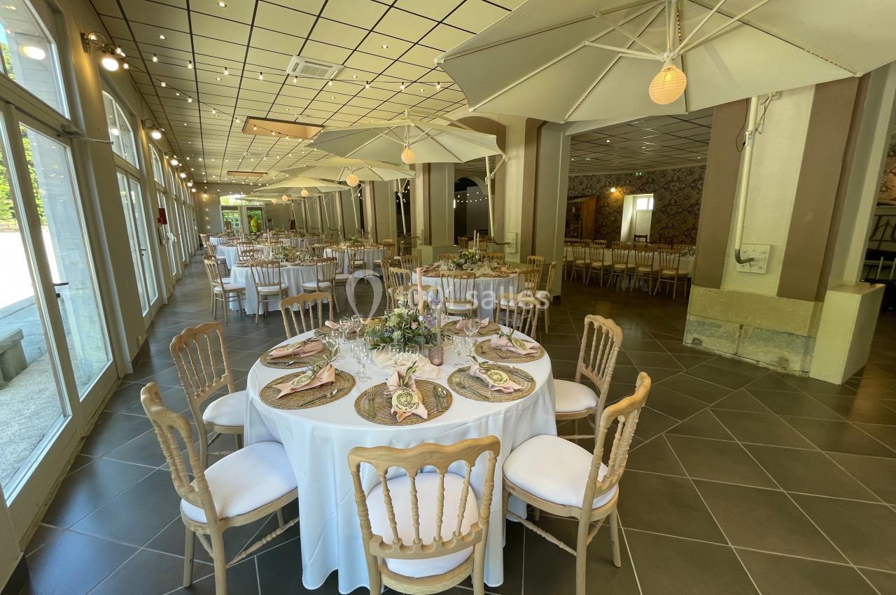 Salle de réception lumineuse avec tables rondes dressées, chaises en bois clair et grands parasols décoratifs.
