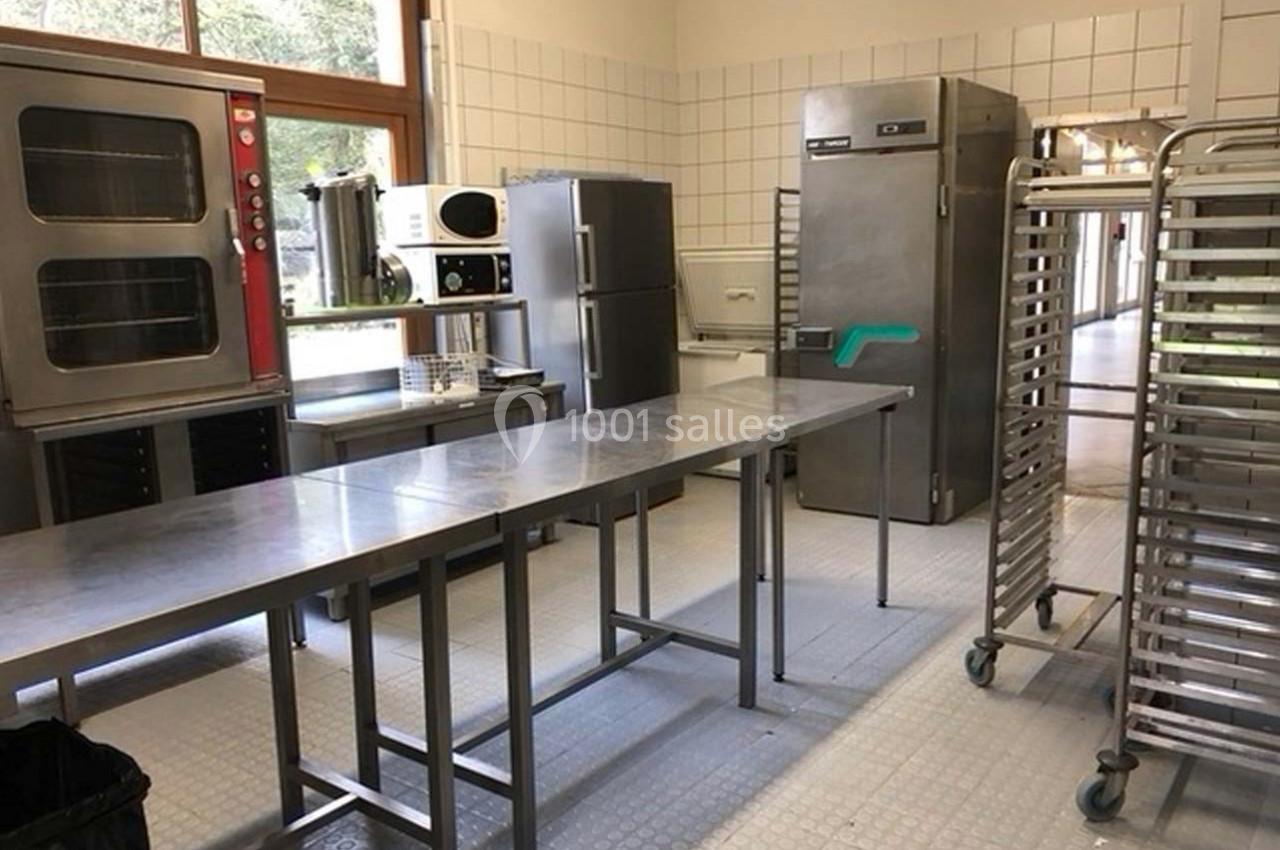 Cuisine professionnelle avec plans de travail en inox, fours, réfrigérateurs et chariots à plateaux sur un sol carrelé.