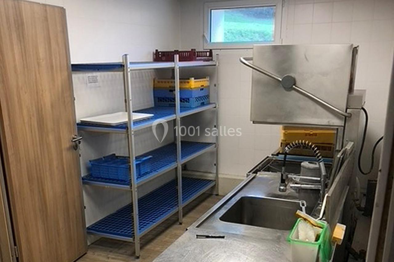 Cuisine professionnelle avec évier inox, lave-vaisselle industriel, étagères métalliques et fenêtre en arrière-plan.