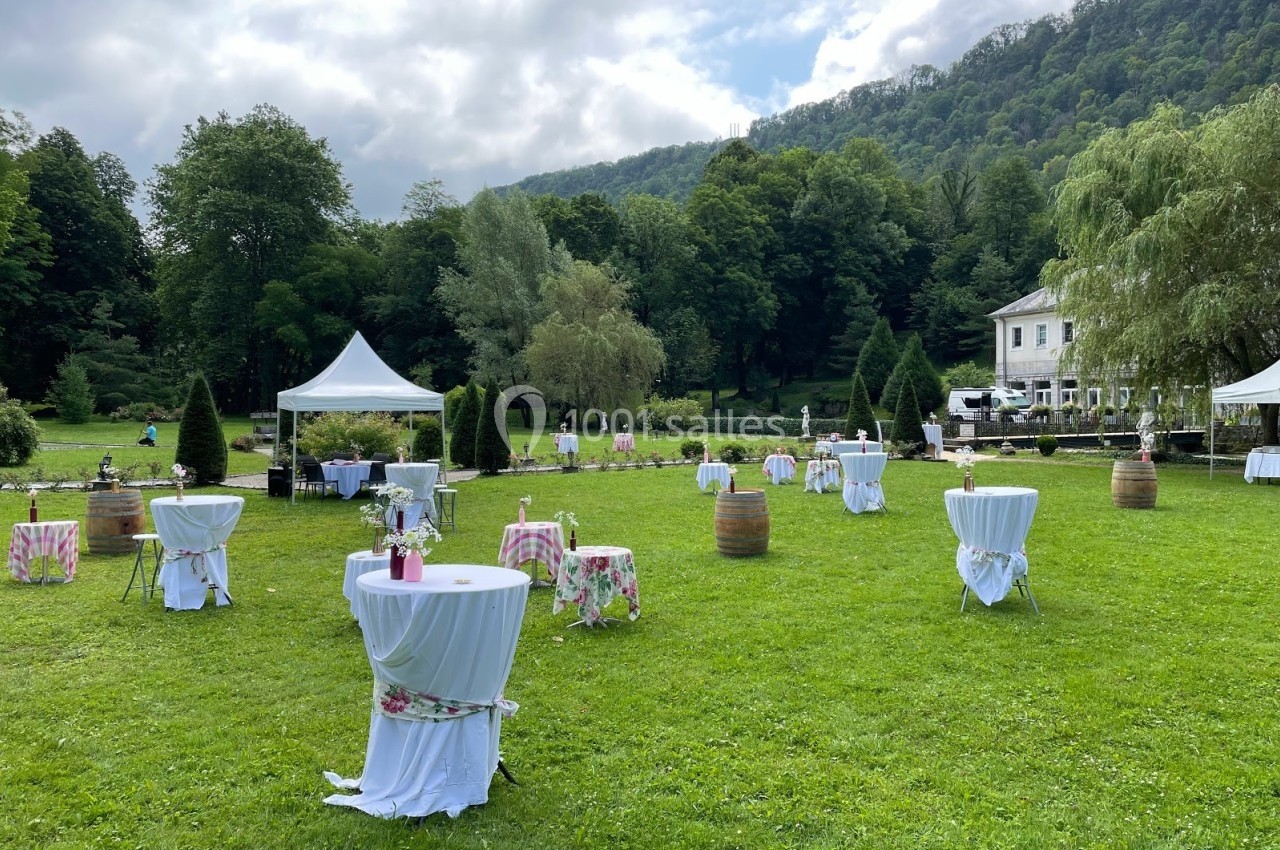 Tables hautes décorées sur une pelouse verdoyante, entourées d'arbres et de montagnes, avec une tente blanche en arrière…