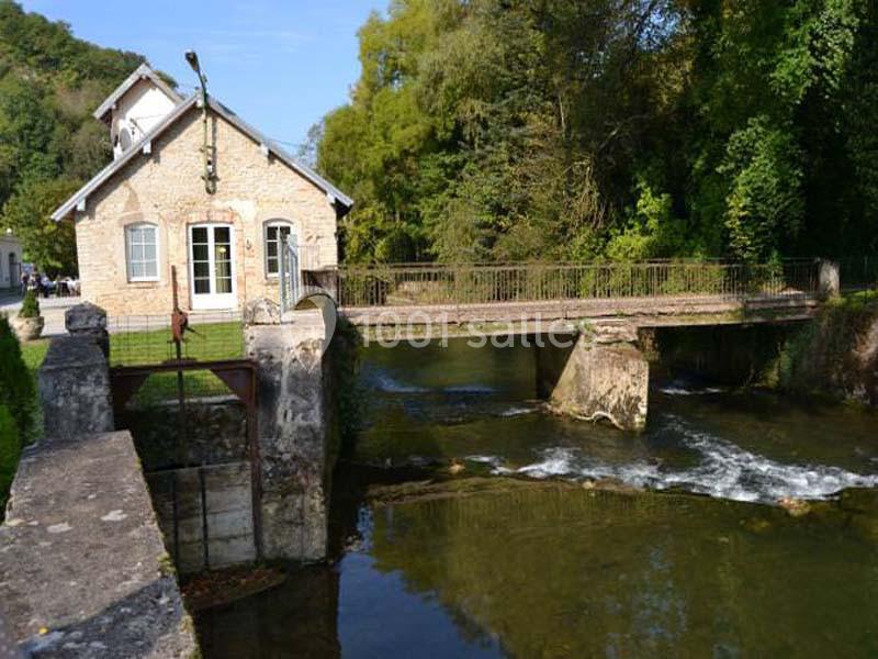 Maison en pierre près d'un petit pont surplombant une rivière avec végétation environnante.