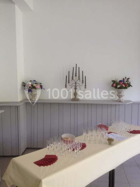 Table dressée avec nappes beige, verres alignés, serviettes rouges, chandeliers et bouquets de fleurs en arrière-plan.