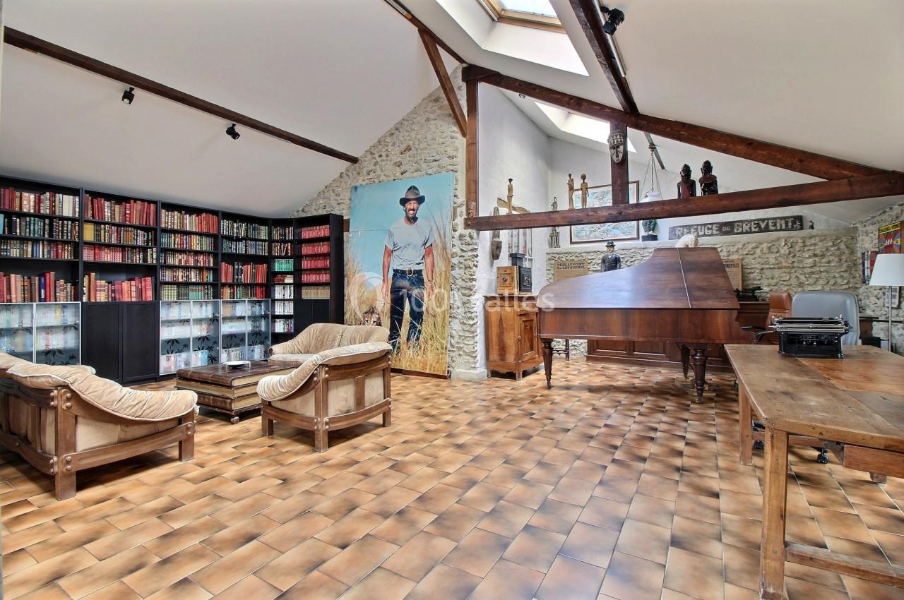 Salle spacieuse avec murs en pierre, bibliothèque, piano à queue, mobilier en bois et grande affiche décorative.
