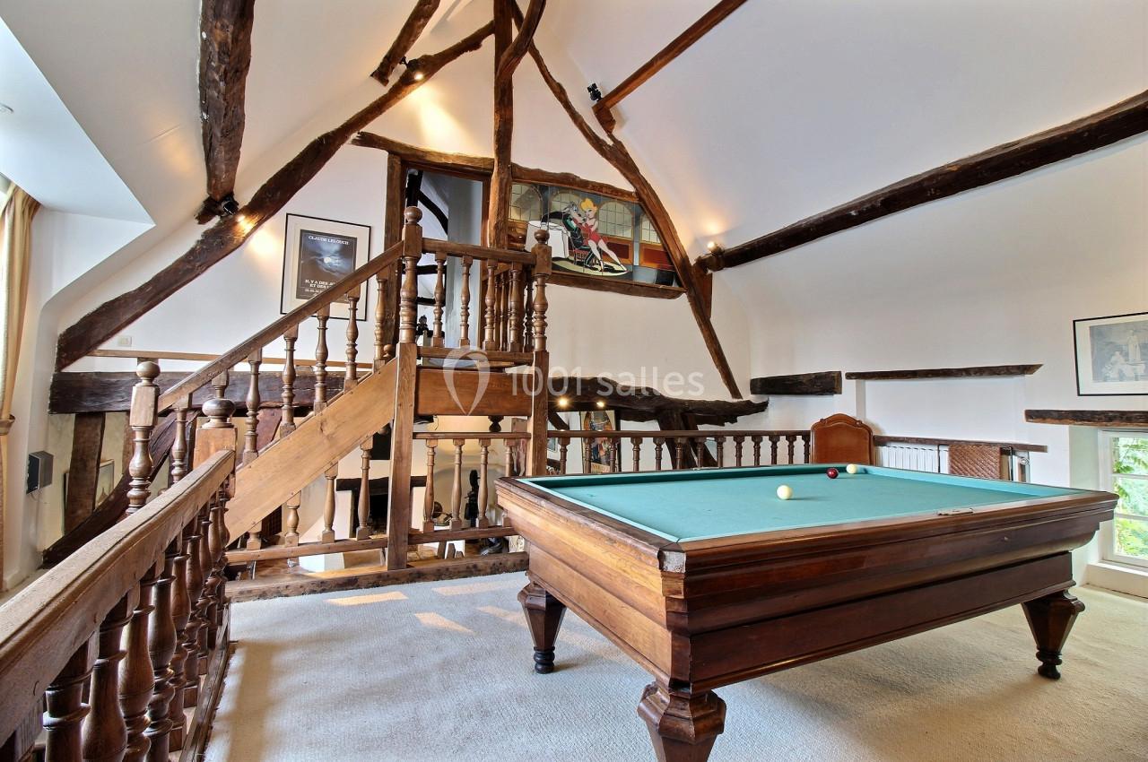 Salle avec poutres apparentes et table de billard en bois, éclairée par des fenêtres et des luminaires muraux.
