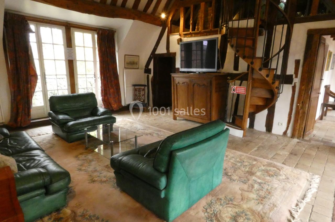 Salon avec poutres apparentes, meubles en bois, canapé vert, tapis beige et escalier en colimaçon en arrière-plan.