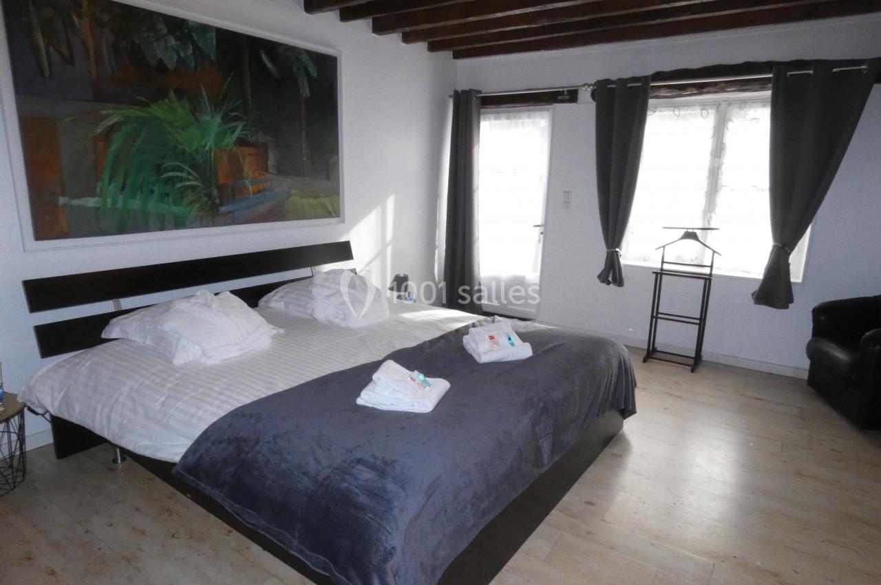 Chambre lumineuse avec un grand lit double, draps blancs, couverture grise, poutres apparentes et grandes fenêtres.