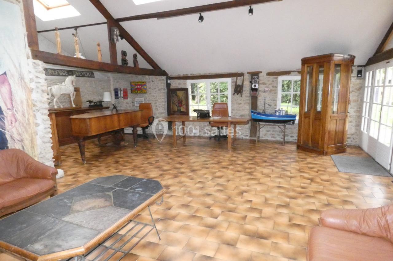 Salle spacieuse avec sol en carrelage, mobilier en bois, poutres apparentes et grandes fenêtres laissant entrer la lumière.