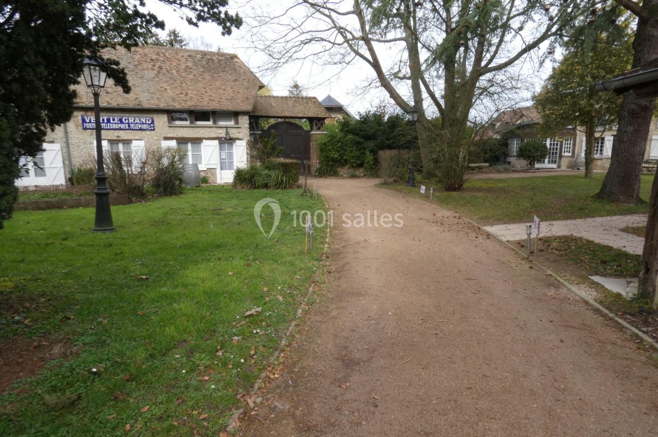 Chemin en gravier bordé de pelouse et d'arbres, menant à des bâtiments en pierre avec toits en tuiles.