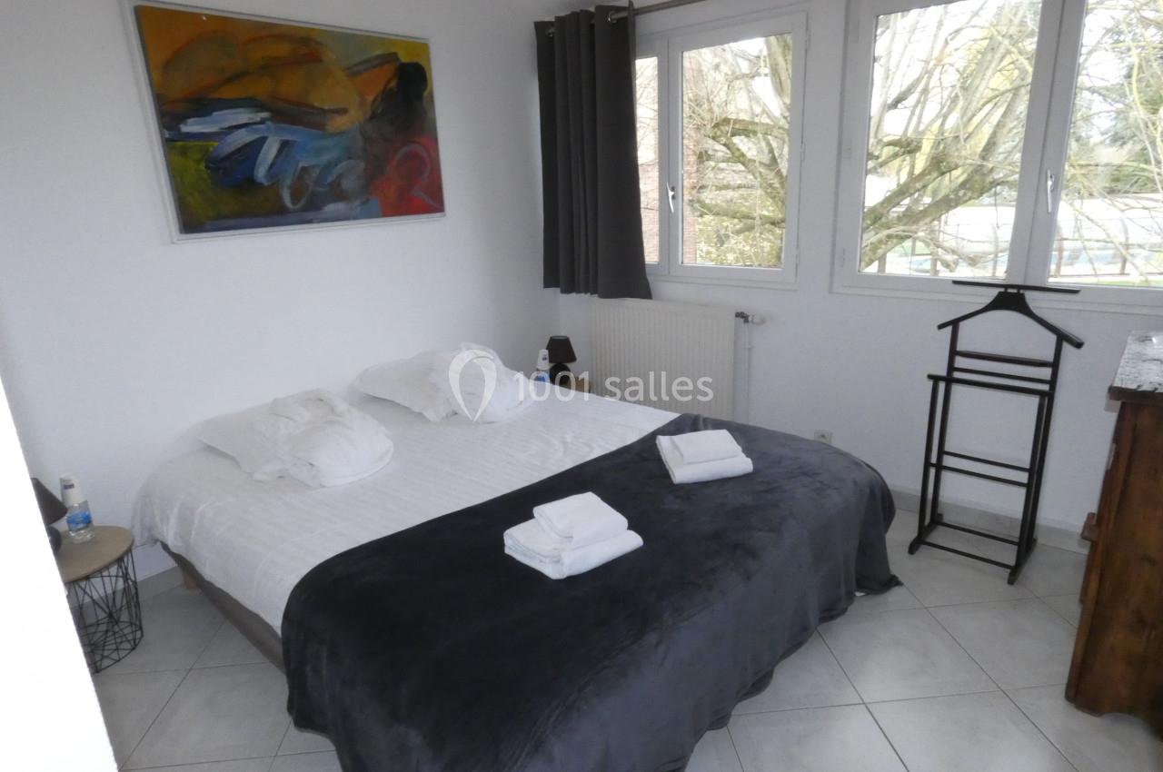 Chambre lumineuse avec un lit double, draps blancs, plaid gris, tableau coloré au mur et vue sur des arbres par la fenêtre.