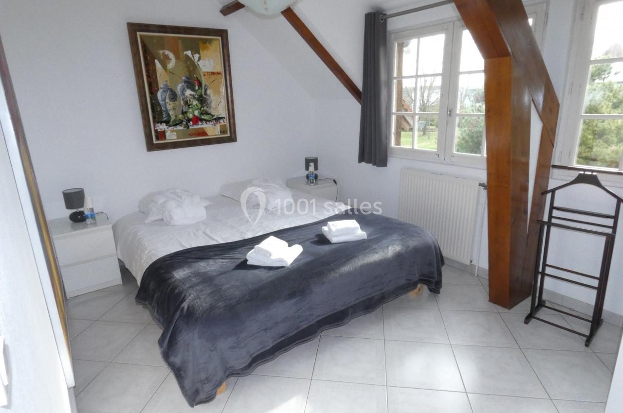 Chambre lumineuse avec lit double, couverture sombre, peignoirs pliés, grande fenêtre et poutres apparentes.