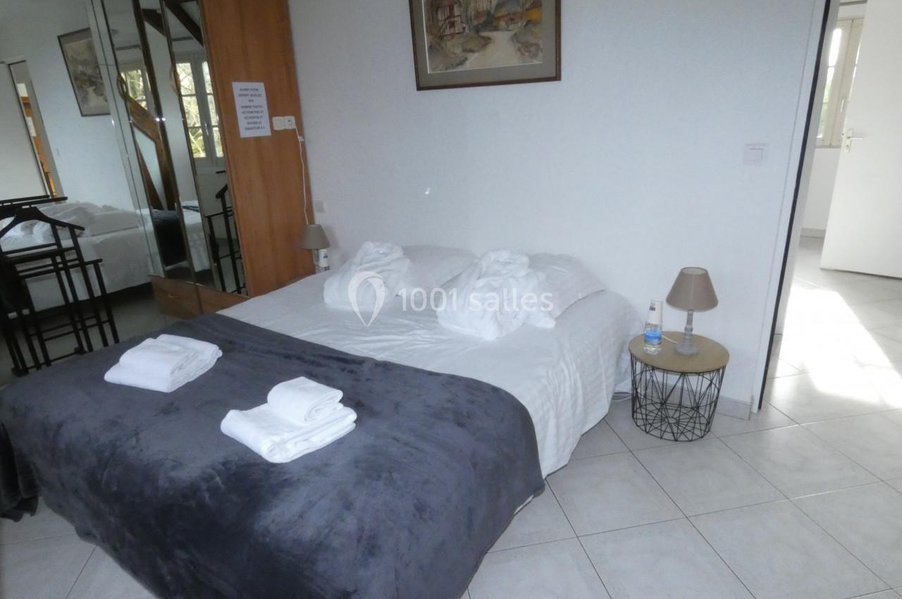 Lit double avec draps blancs, plaid gris, peignoirs et serviettes pliées dans une chambre lumineuse au sol carrelé.