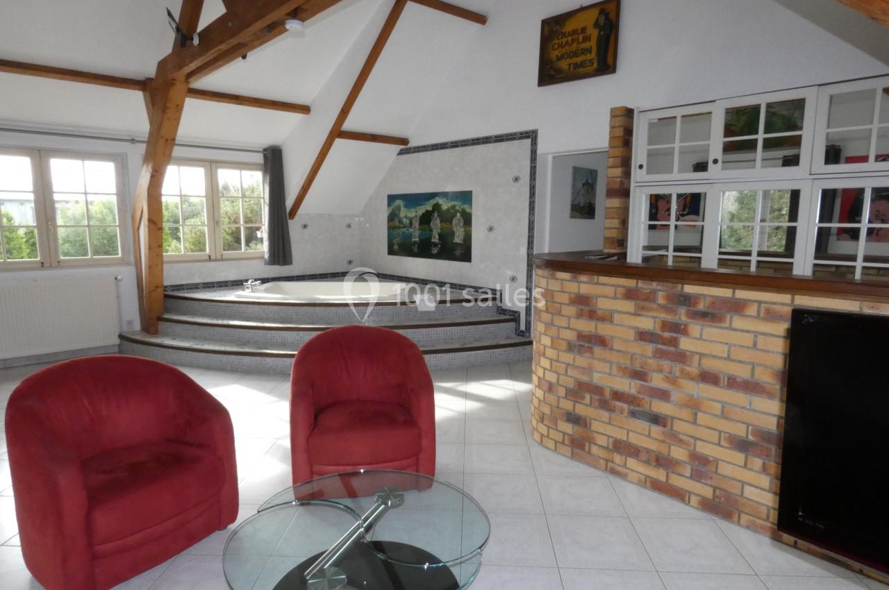 Salle lumineuse avec jacuzzi sur estrade, fauteuils rouges, table basse en verre et bar en briques.