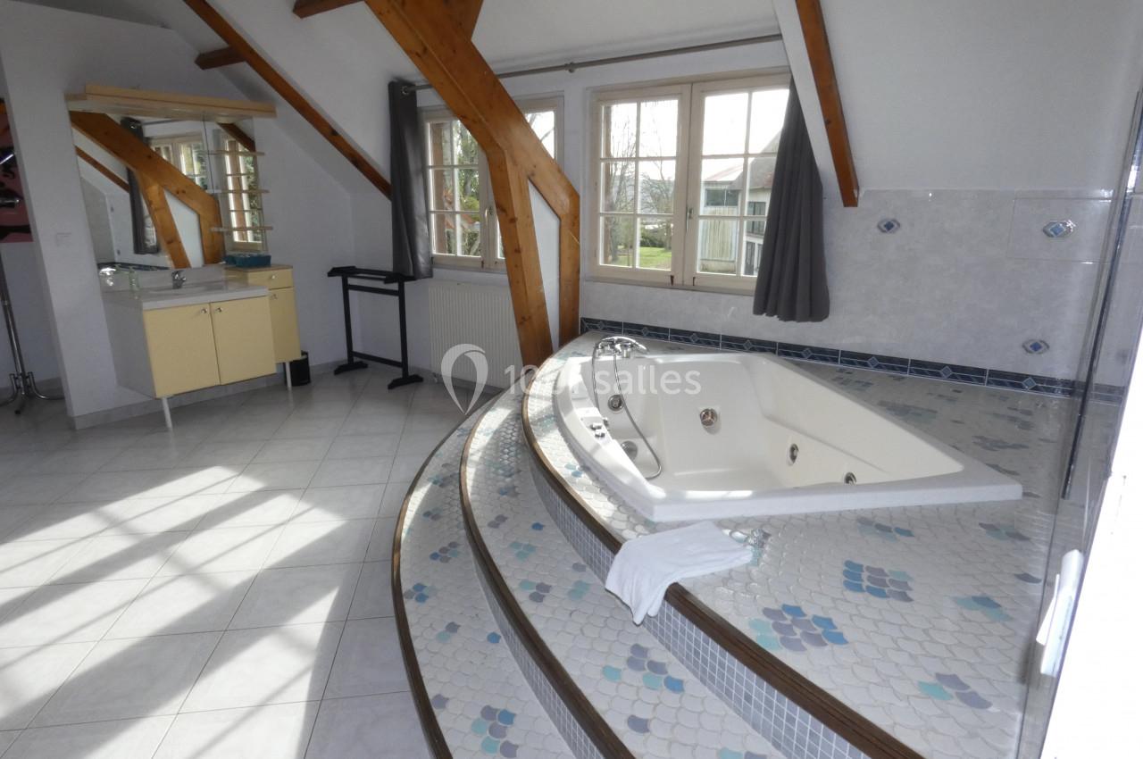 Salle de bain lumineuse avec baignoire encastrée sur estrade carrelée, poutres apparentes et grande fenêtre.