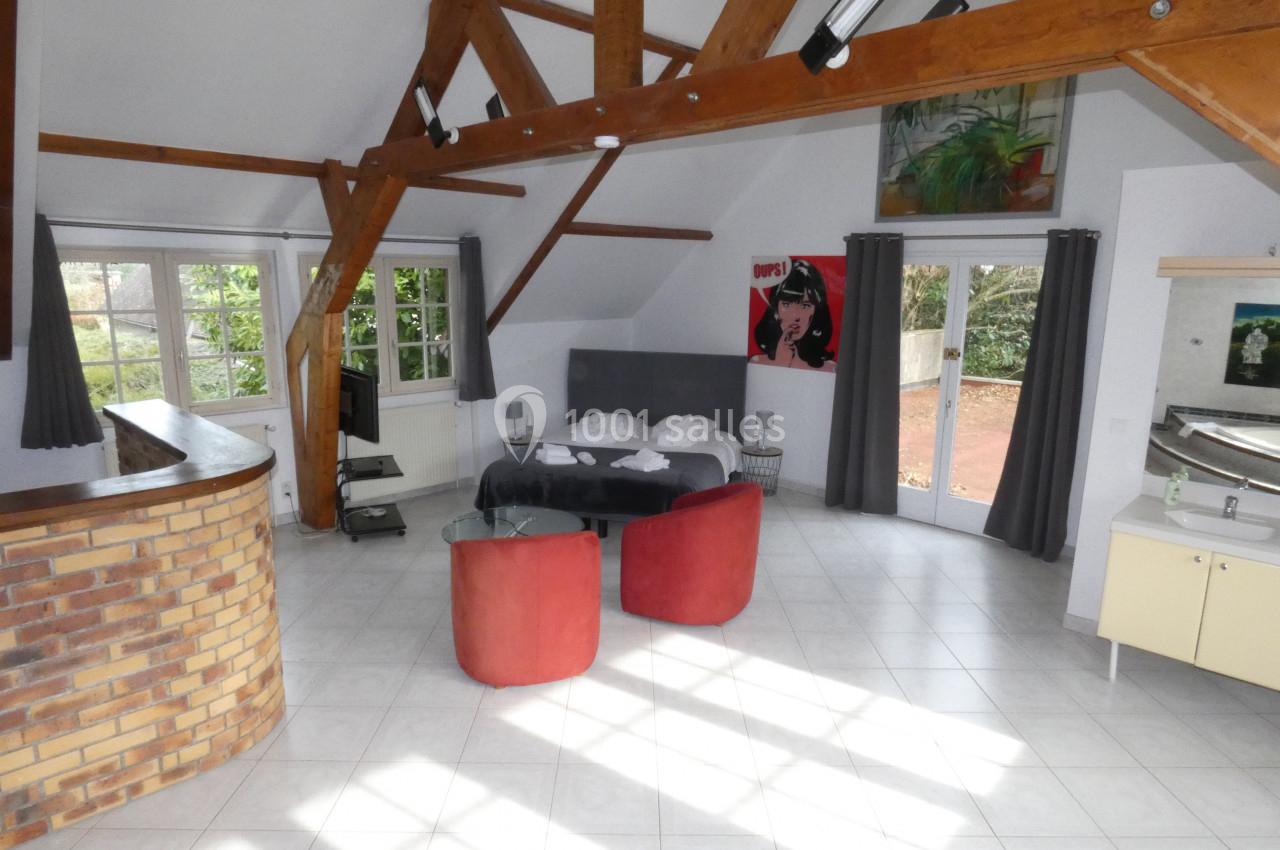 Grande pièce lumineuse avec poutres apparentes, lit double, fauteuils rouges, bar en briques et grandes fenêtres.