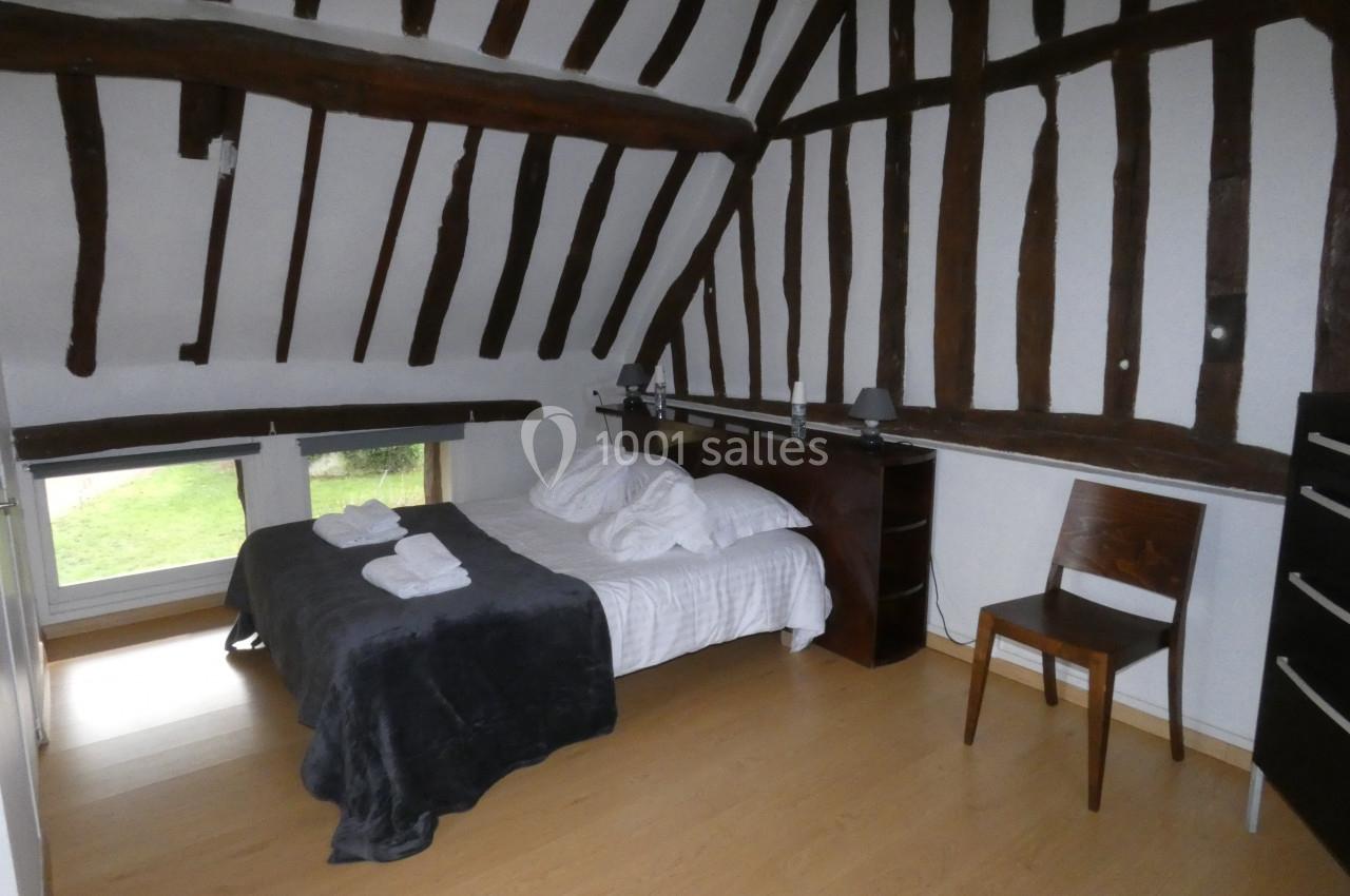 Chambre avec poutres apparentes, lit double, chaise en bois, et fenêtre donnant sur un espace extérieur verdoyant.