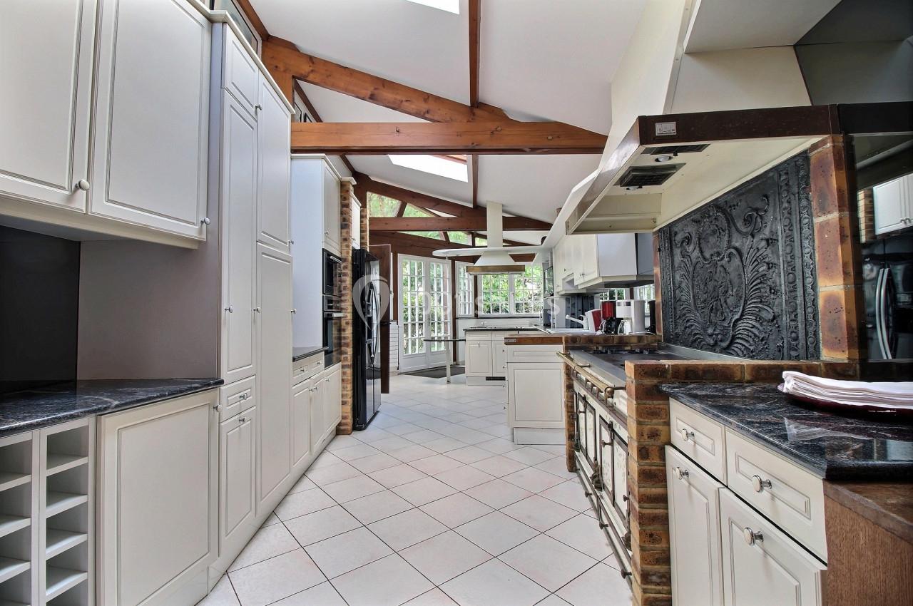 Cuisine spacieuse avec poutres apparentes, meubles blancs, plan de travail sombre et vue sur un jardin à travers de grandes…