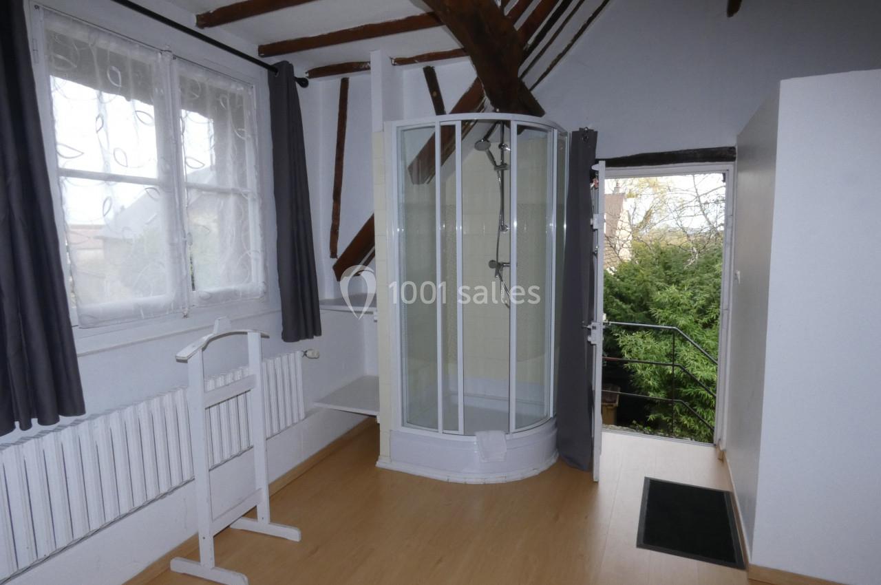 Chambre mansardée avec parquet, douche d'angle, fenêtre avec rideaux gris et vue sur un espace extérieur verdoyant.