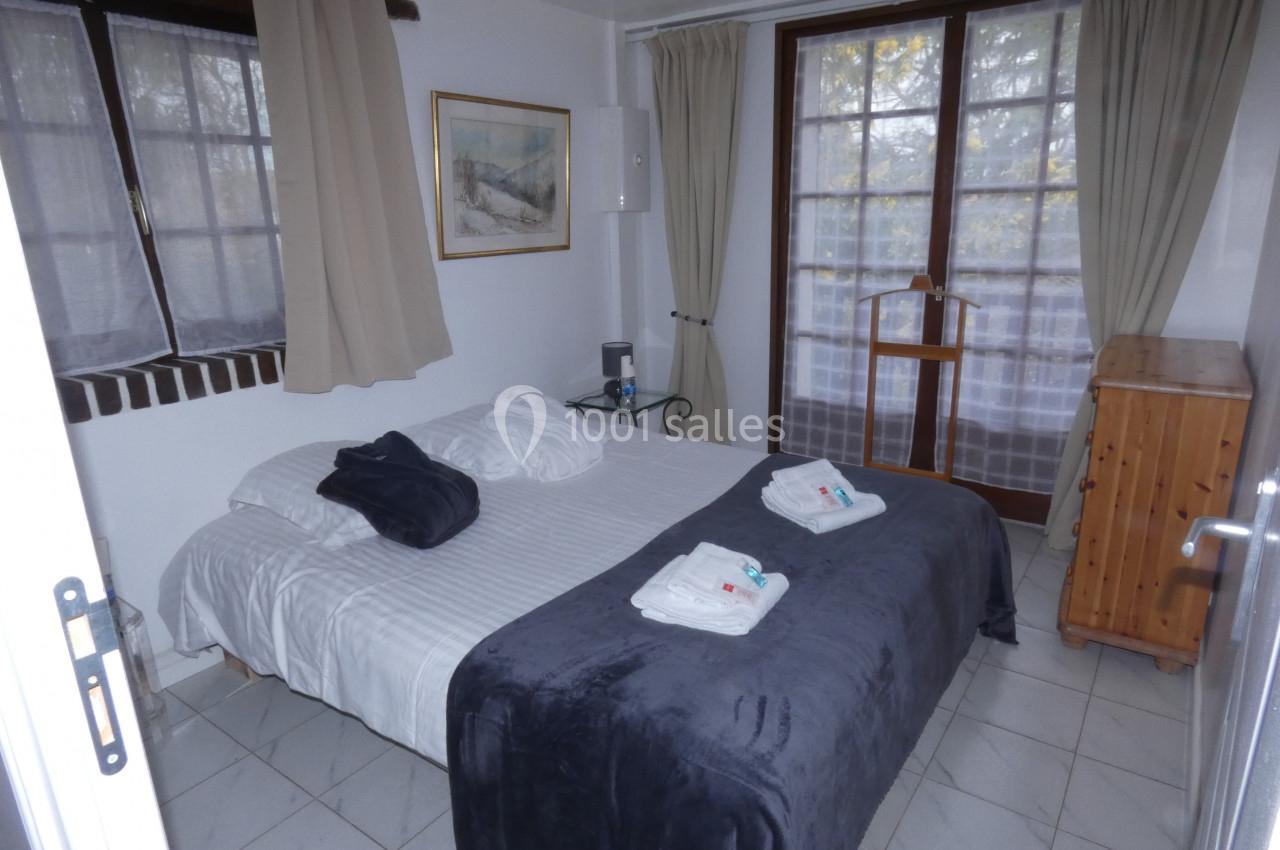 Chambre avec lit double, couverture grise, fenêtres avec rideaux clairs et mobilier en bois.