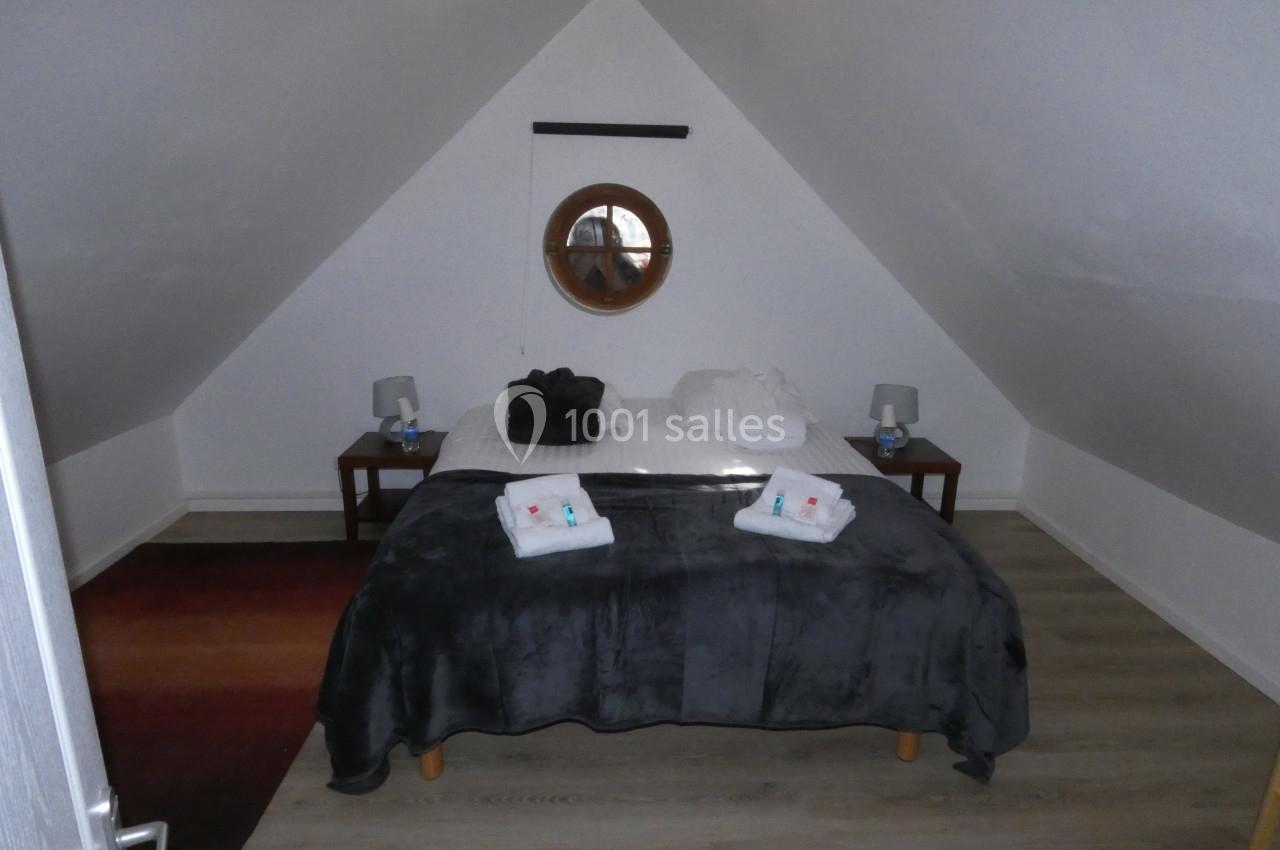 Chambre mansardée avec un lit double, draps pliés, tables de chevet avec lampes et un miroir rond au mur.