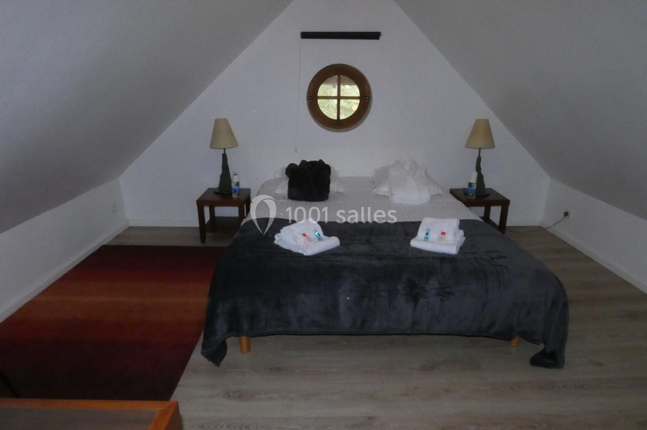 Chambre mansardée avec lit double, deux lampes de chevet, fenêtre ronde et sol en parquet clair.