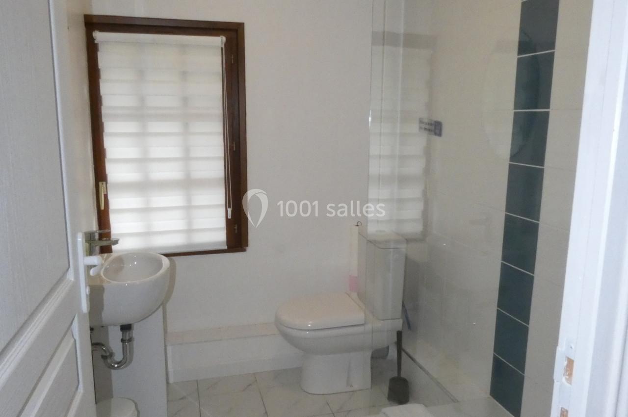 Salle de bain avec douche à l'italienne, lavabo, toilettes, carrelage blanc et fenêtre avec store.