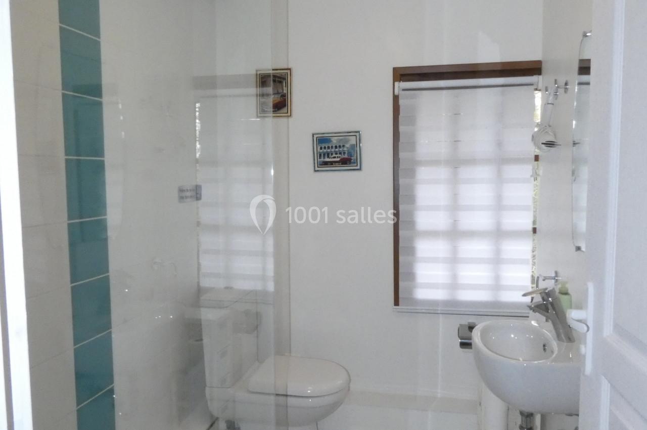 Salle de bain lumineuse avec douche à l'italienne, lavabo, toilettes et fenêtre avec store semi-ouvert.
