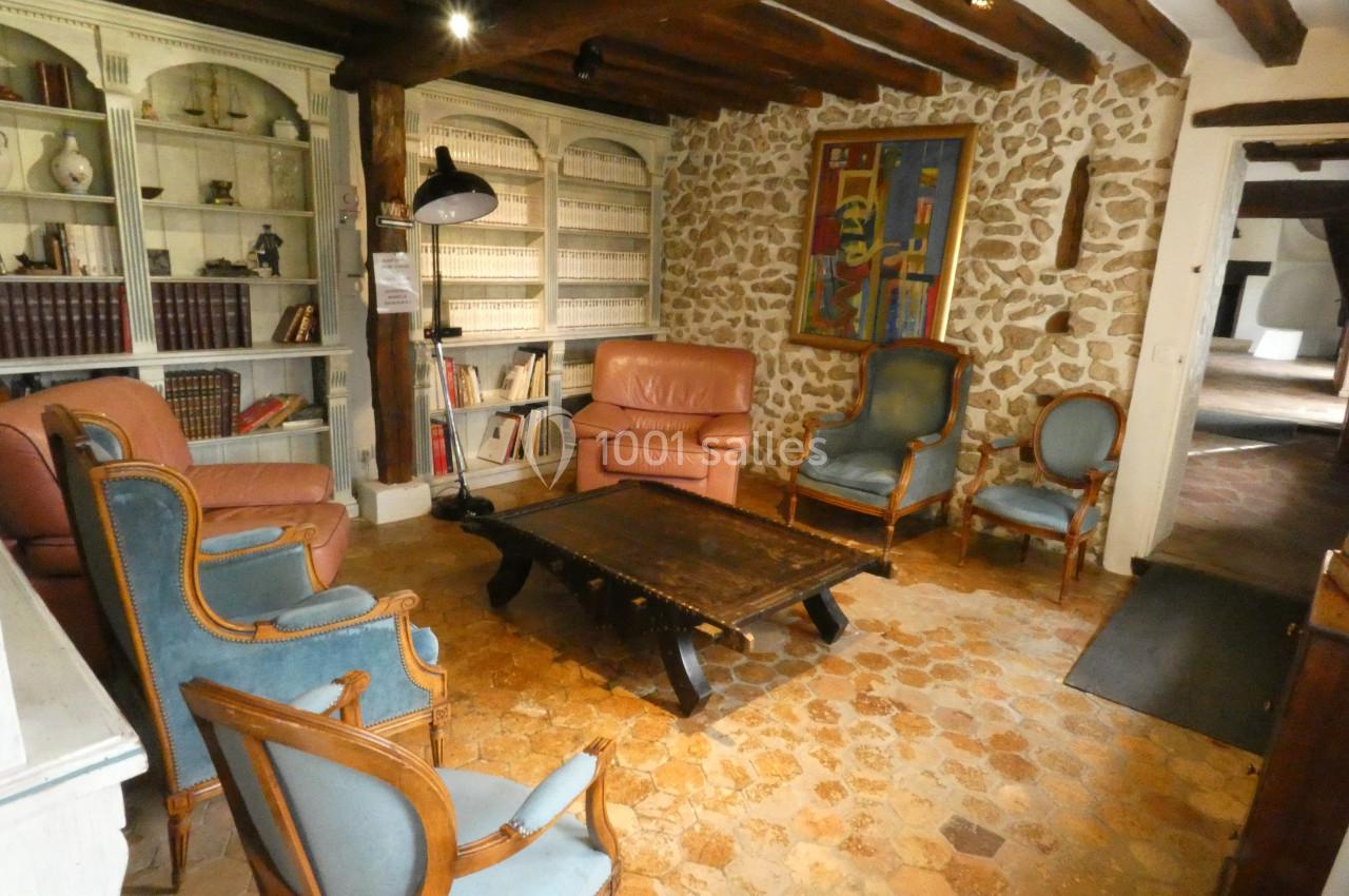 Salon rustique avec murs en pierre, poutres apparentes, bibliothèque, fauteuils anciens et table basse en bois.