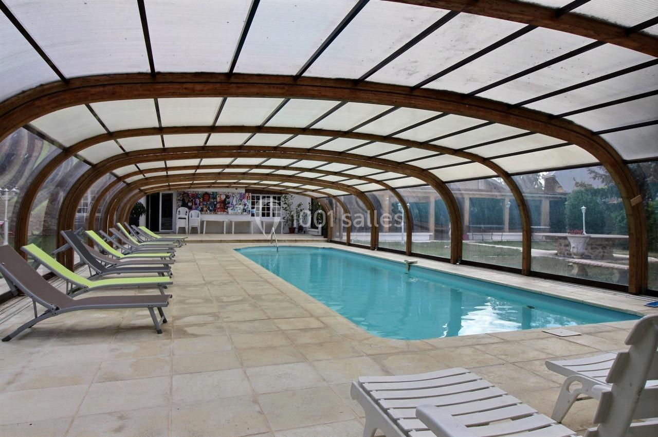 Piscine intérieure couverte avec structure en bois, entourée de chaises longues sur un sol carrelé.